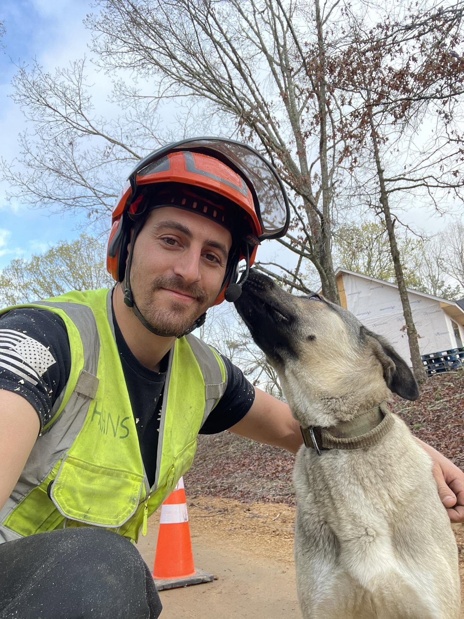 A Hahn's Tree Service crew member in helmet and hi-vis vest crouched beside a friendly dog on a job site