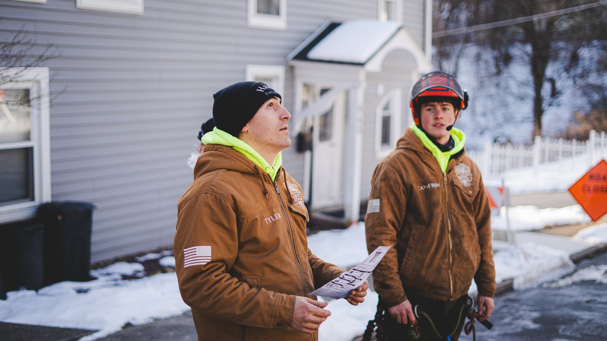 Two crew members outside a house looking up at a tree with a work order in hand, road closed sign visible behind them
