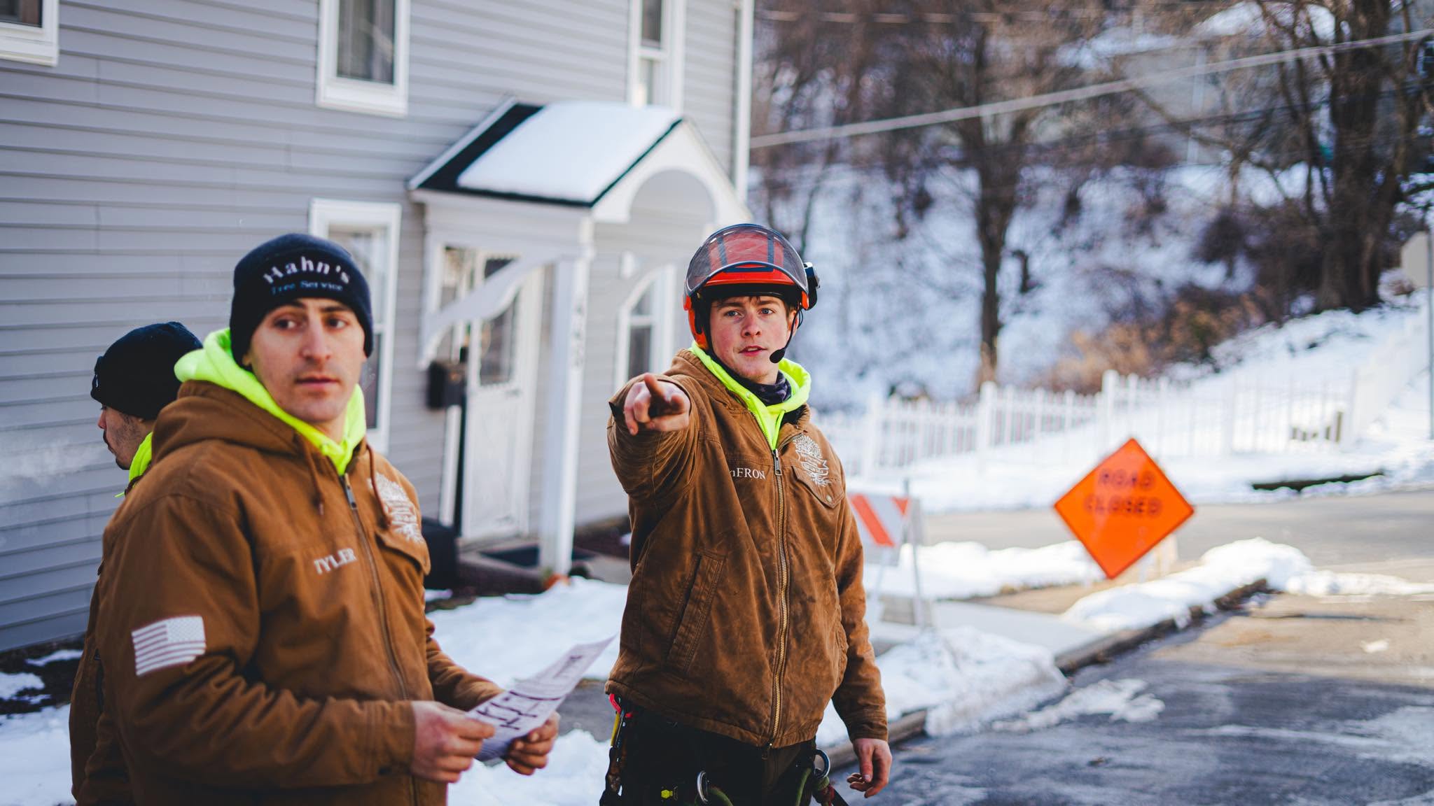 Two Hahn's Tree Service crew members in Carhartt jackets on a winter street with a road closed sign, one pointing toward the job